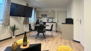 A modern living room with a black sofa, a dining table with chairs, a TV mounted on the wall, and a kitchen area in the background.