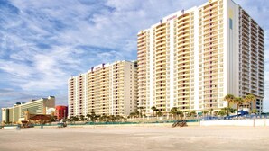 Un grand hôtel à plusieurs étages, doté de nombreux balcons, situé près d’une plage bordée de palmiers, sous un ciel dégagé.