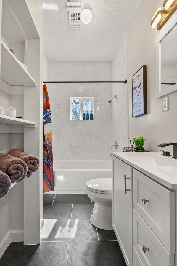 Un baño moderno con bañera, inodoro y zona de ducha. Hay toallas en un estante, un cuadro en la pared y una planta en el mostrador del lavabo.