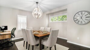 A dining room with a wooden table set for six, six grey upholstered chairs, a chandelier, a large wall clock, and a window with a view of greenery. - Charming 3-bedroom house close to Promedica and UToledo (Toledo)