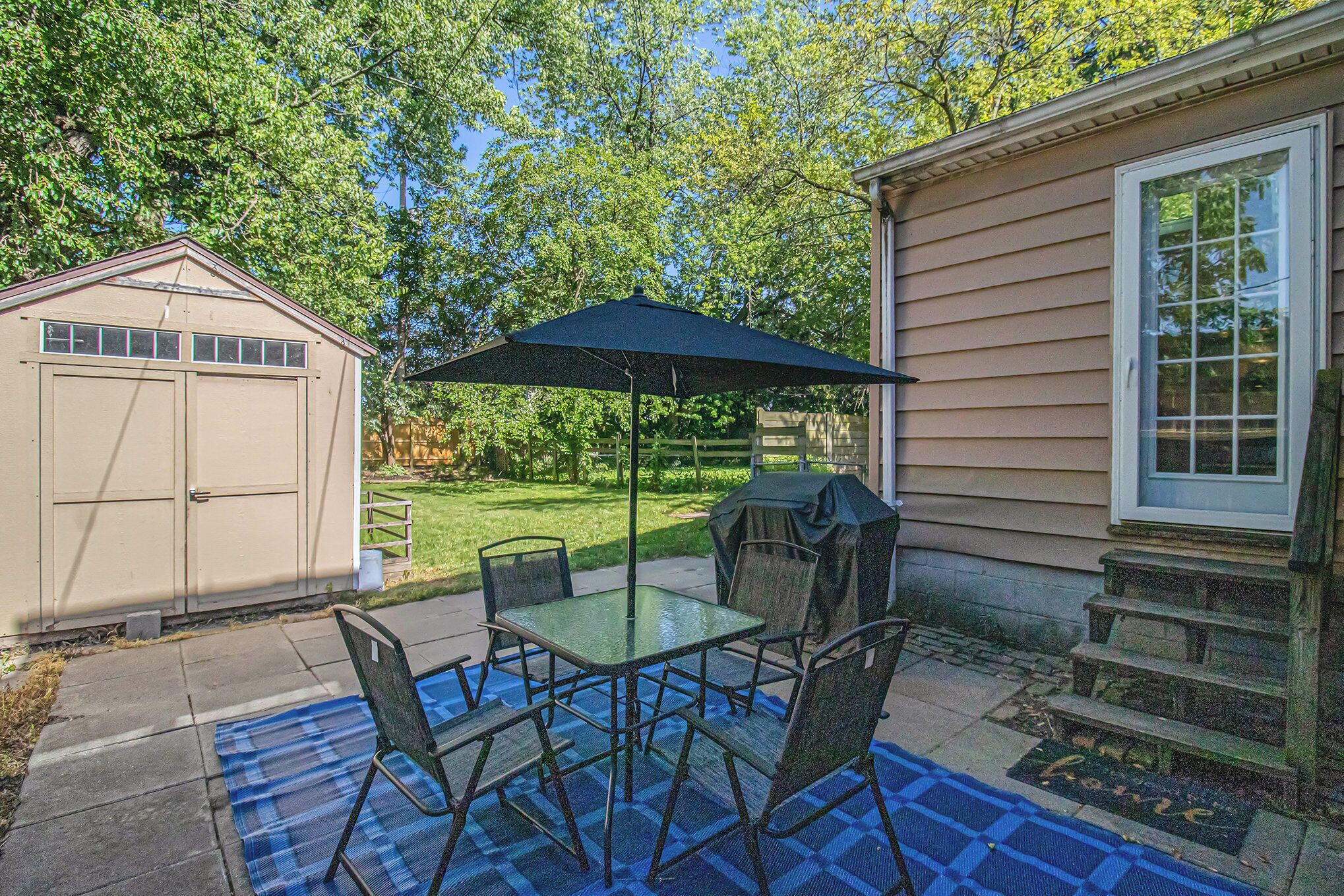 Une terrasse avec une table et des chaises en verre, un tapis à carreaux bleus et un parasol noir.