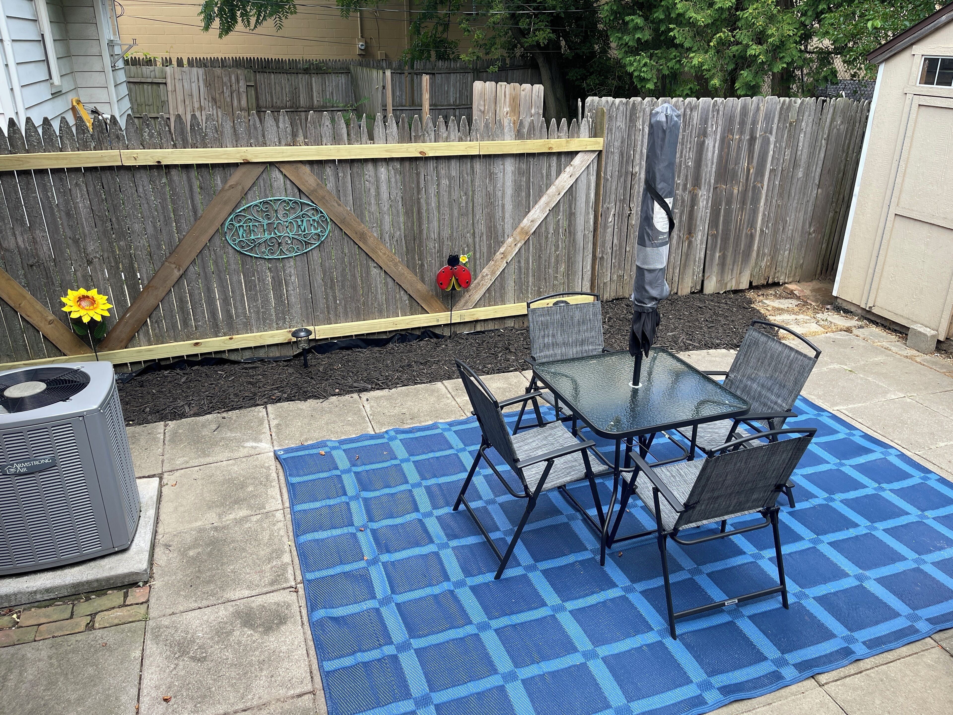 A backyard patio with a glass table and chairs, a blue checkered mat, and a welcome sign on a wooden fence.