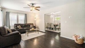 A modern living room with dark wood flooring, a ceiling fan, a dining area with a chandelier, and a view of the outdoors. - Charming 3-bedroom house close to Promedica and UToledo (Toledo)