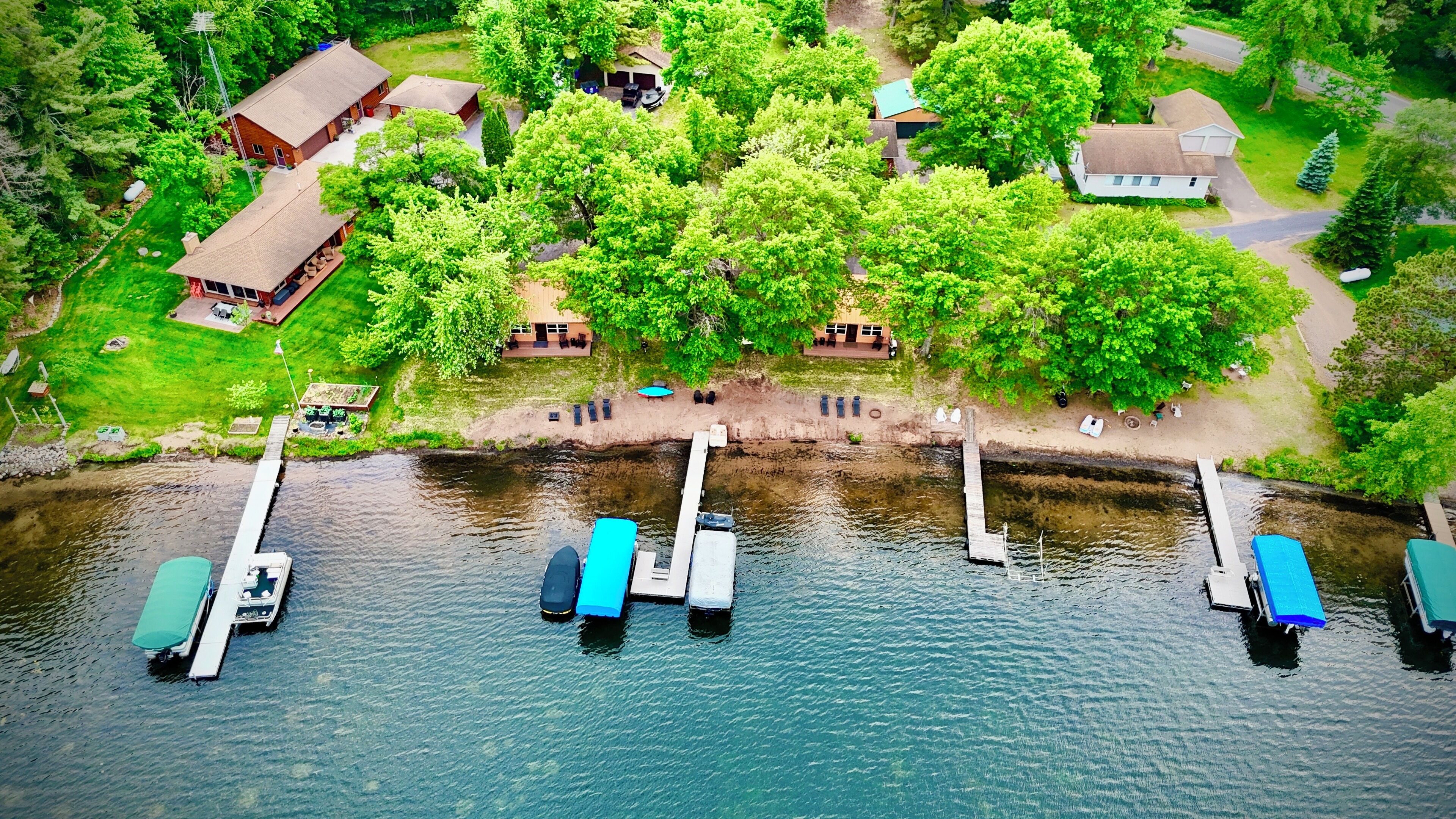 An aerial view of a lakeside property with docked boats, a small dock, and several buildings surrounded by lush greenery.