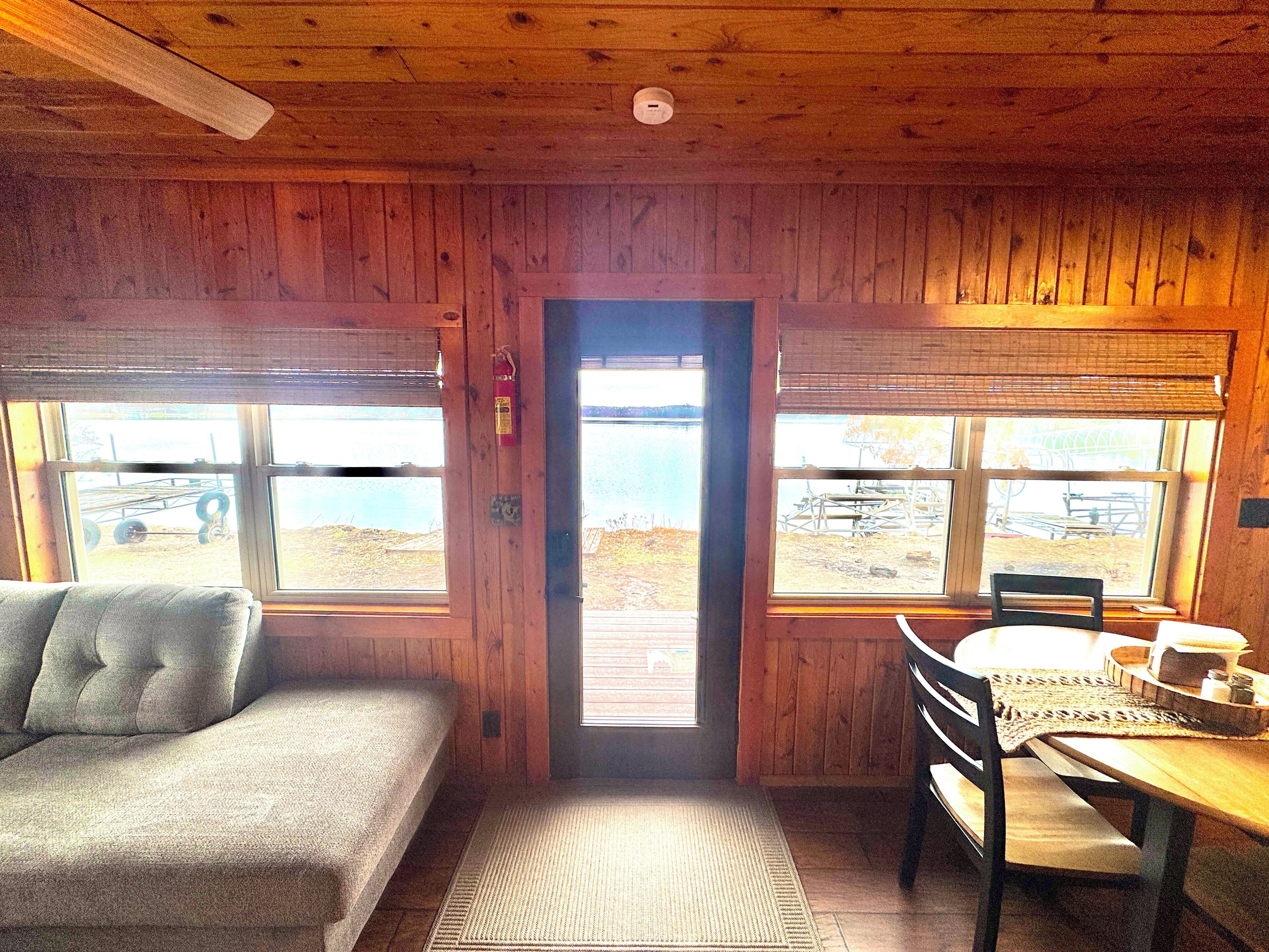A cabin interior with a sofa, a dining table with chairs, and large windows offering a view of the outdoors.
