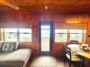 A cabin interior with a sofa, a dining table with chairs, and large windows offering a view of the outdoors.