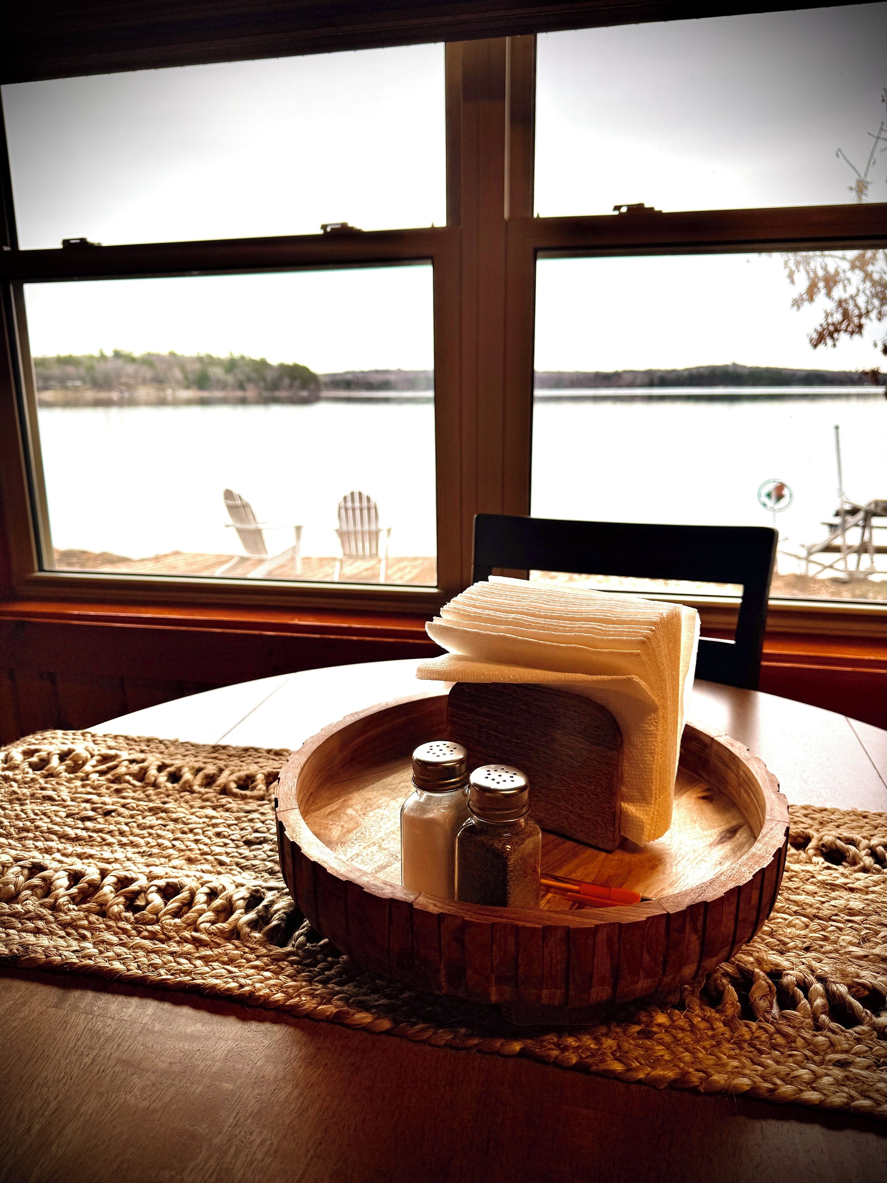 Une table en bois, sur laquelle est posée une nappe en fibres tressées, une pile de serviettes blanches, deux salières et deux poivrières, et une chaise.