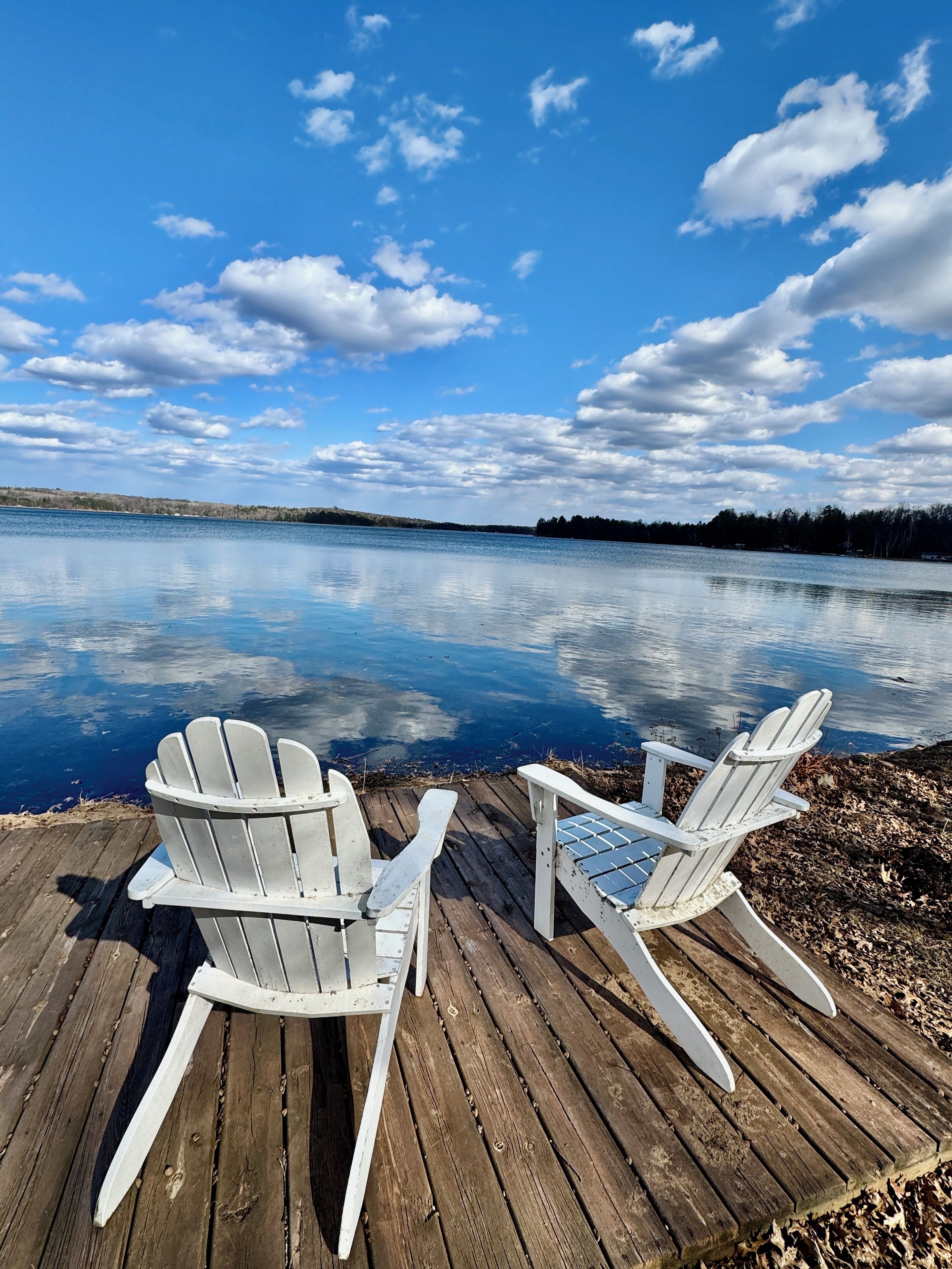Two white Adirondack chairs on a wooden dock by a calm lake with a clear sky.