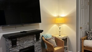 Un salón con chimenea de piedra, un televisor de pantalla plana montado encima, una silla de mimbre con una manta, una mesita con una lámpara y una puerta al fondo.