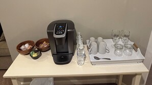 Una mesa con cafetera, botellas de agua, tazas y una bandeja con vasos. En la pared hay carteles enmarcados y un cubo de basura debajo de la mesa.
