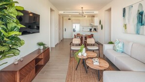 Ein modernes Wohnzimmer mit Sofa, Couchtisch und Fernseher. Es gibt einen Essbereich mit Stühlen und einem Tisch. Die Zimmer ist mit einer großen Pflanze und einem Bild an der Wand ausgestattet.