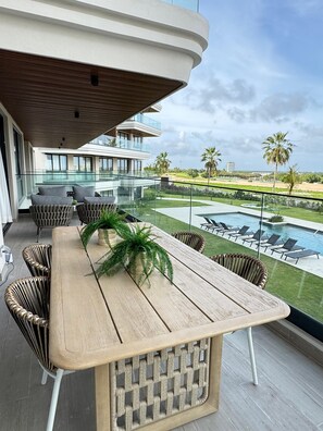 A modern outdoor dining area with a wooden table, wicker chairs, and a view of a swimming pool and palm trees.