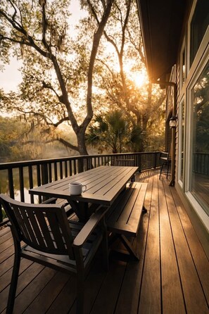 A wooden deck with a table and benches, overlooking a body of water and trees, with a building on one side.