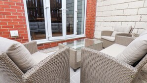 Un patio con mobiliario de mimbre y una mesa de vidrio, junto a un muro de ladrillo y una puerta corrediza de vidrio.