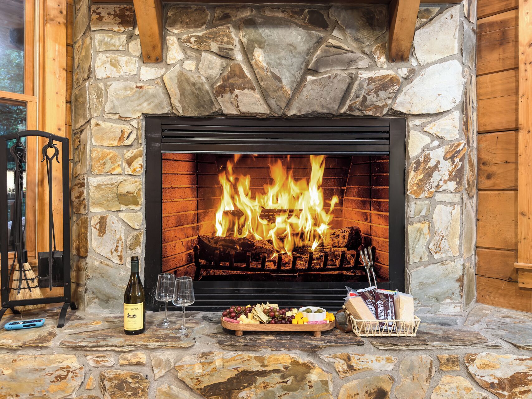 A stone fireplace with a fire, a bottle of wine, glasses, and a tray of snacks.