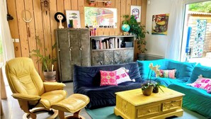 A cozy living room with a yellow leather chair, a dark blue sofa, and a yellow coffee table. There is a bookshelf filled with books and decorative items, and a large window with a view of greenery outside. - House with pool near Montpellier Plage July 12-August 2 (Le Crès)
