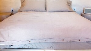 Una cama bien hecha con edredón y almohadas de color claro, una mesita de noche con lámpara y un enchufe en la pared.