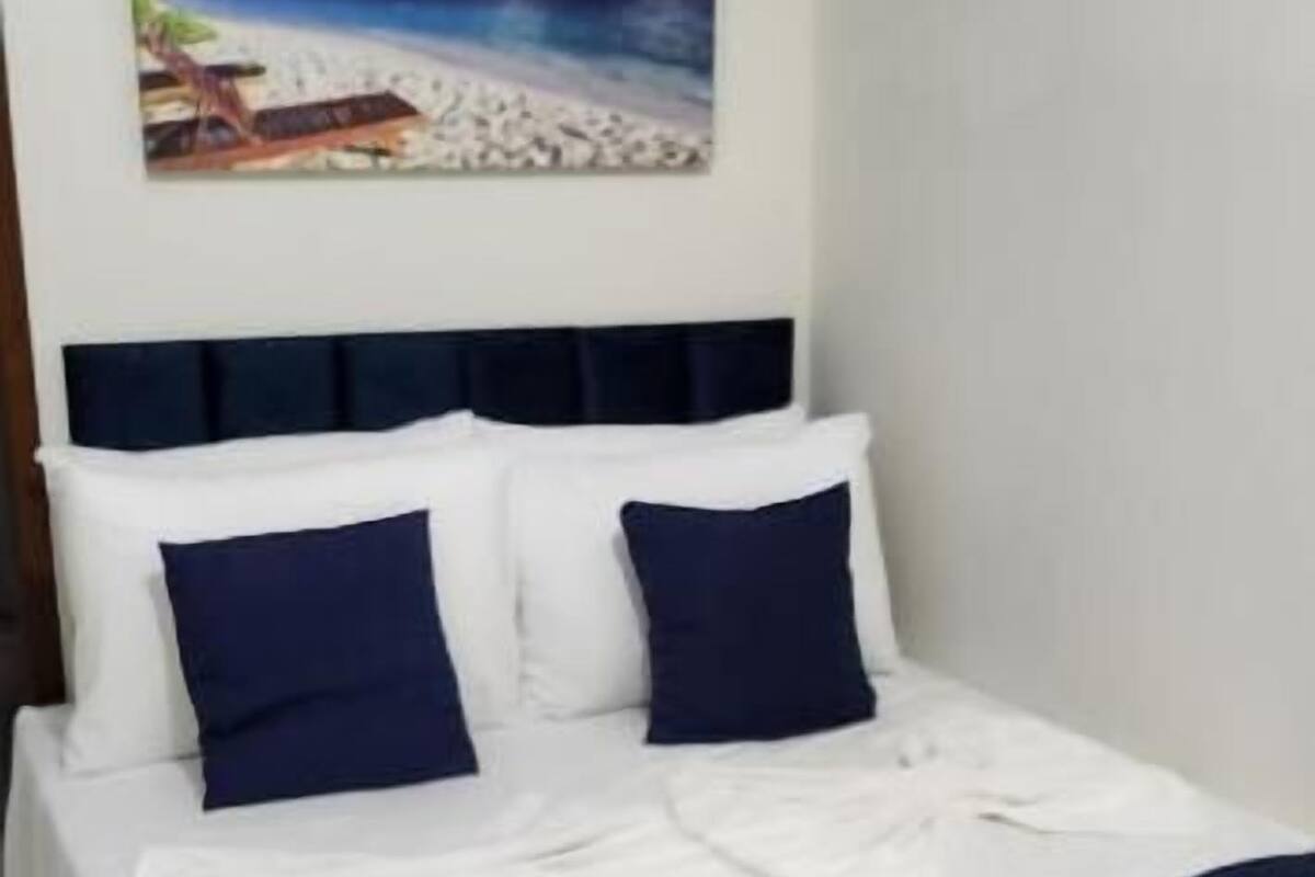 A neatly made bed with white and navy blue bedding, a headboard, and a painting of a beach scene above it.