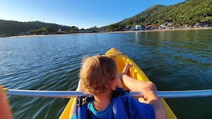 A person in a yellow kayak on calm water with a hilly landscape and buildings in the background. - Pousada Enseada São Miguel (Penha)