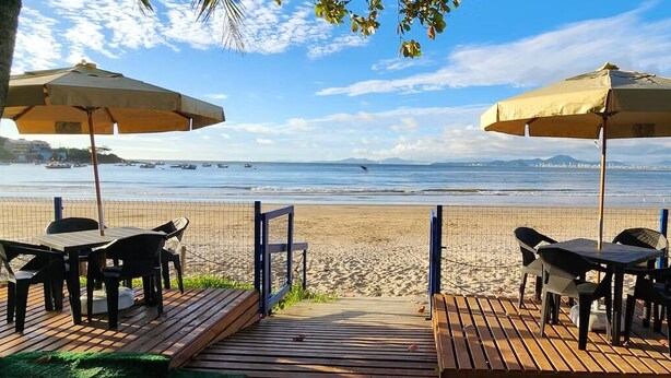 A beachside restaurant with outdoor seating, a wooden deck, and a view of the ocean and city skyline. - Pousada Enseada São Miguel (Penha)