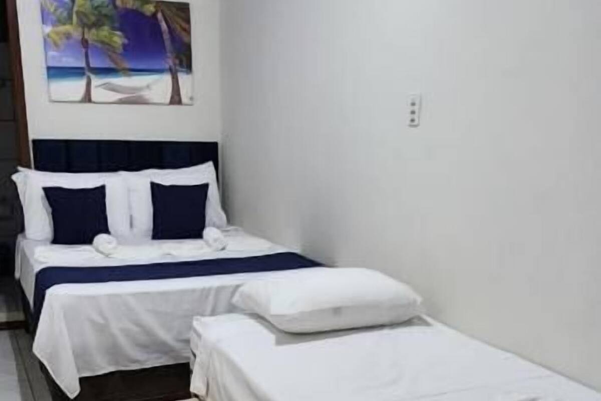 A hotel room with a bed, a bench, a painting, and an air conditioning unit.