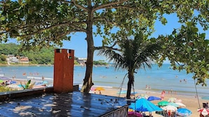 A beach with umbrellas, a palm tree, and a brick structure. - Pousada Enseada São Miguel (Penha)