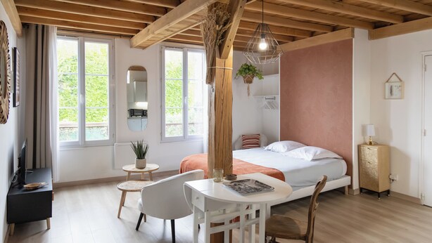 Une chambre à coucher comprenant un lit, une table de chevet, une chaise et un bureau avec une autre chaise. On y trouve une fenêtre avec des rideaux, une plante suspendue et un plafond en bois.