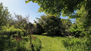 Un jardin luxuriant, avec une grande variété de plantes et de fleurs, un sentier bordé de gazon et des arbres qui offrent de l’ombre.