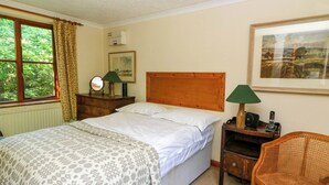 Une chambre à coucher avec un lit, une table de chevet en bois surmontée d’une lampe, un fauteuil en osier et une fenêtre avec des rideaux.
