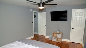 Ein Schlafzimmer mit einem Bett, einem Deckenventilator, einem an der Wand montierten Fernseher und einer Tür zu einem weiteren Raum.