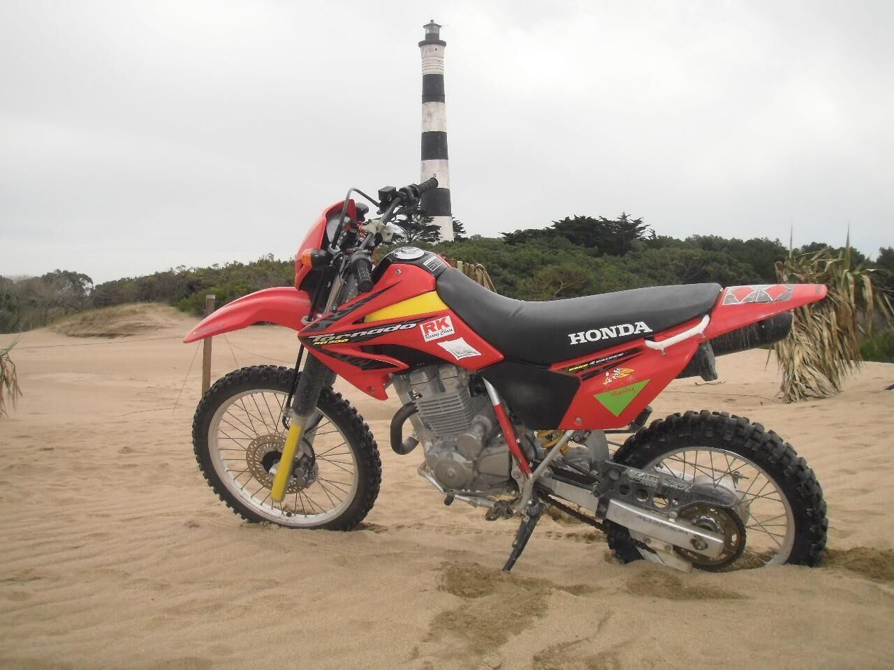 Ein rotes Honda-Dirtbike steht auf einem sandigen Strand, im Hintergrund ein Leuchtturm.
