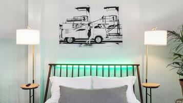 Habitació d'hotel amb llit, dues taules de nit, una làmpada penjada a la paret, un panell de capçalera il·luminat de verd i una obra d'art en blanc i negre que representa un camió de remolc.