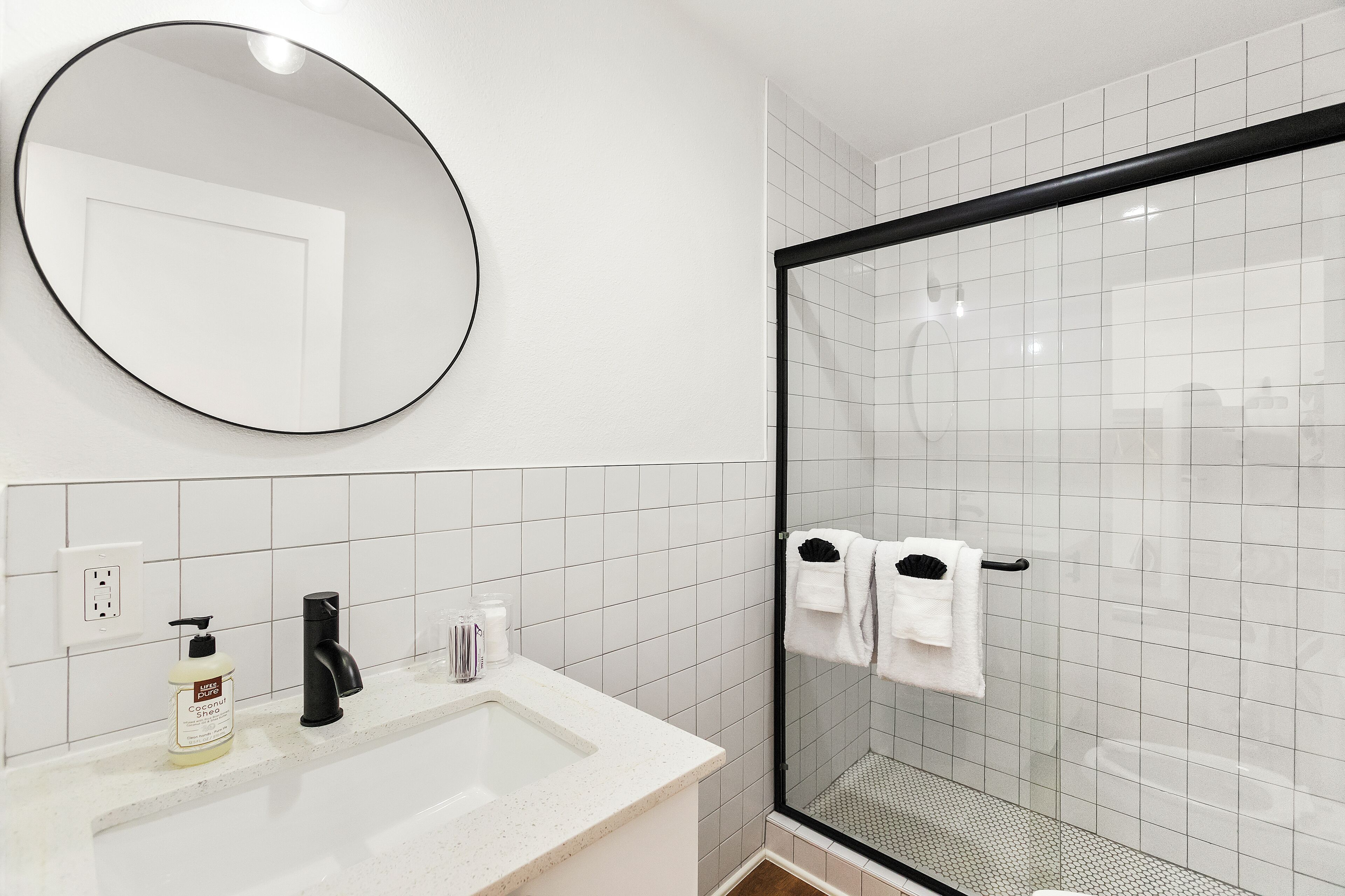 Een badkamer met een witte wastafel, een zwarte kraan, een ronde spiegel en een douchecabine van glas.