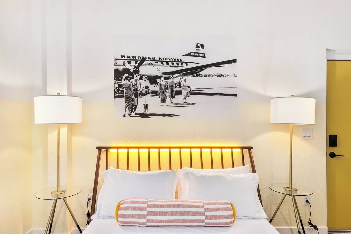 A hotel room with a bed, two bedside lamps, a wall-mounted picture of Hawaiian Airlines, and a yellow door.