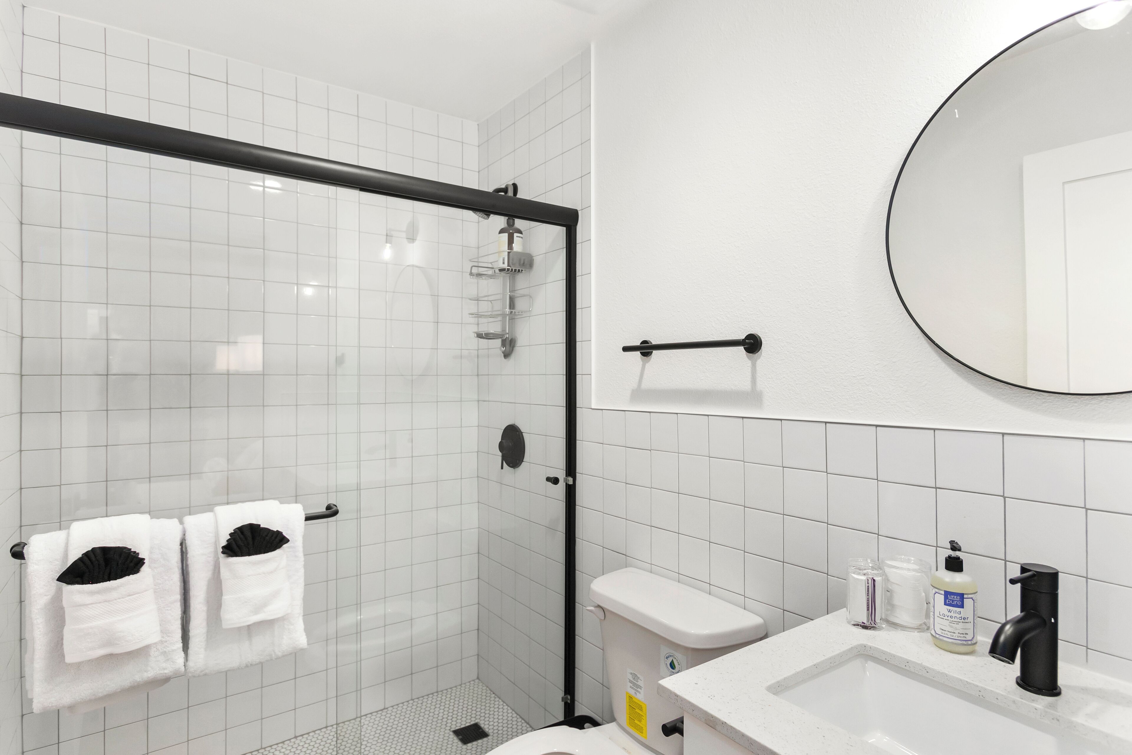 A modern bathroom with a glass-enclosed shower, white tiles, a black towel rack, a white sink with a black faucet, and a round mirror.
