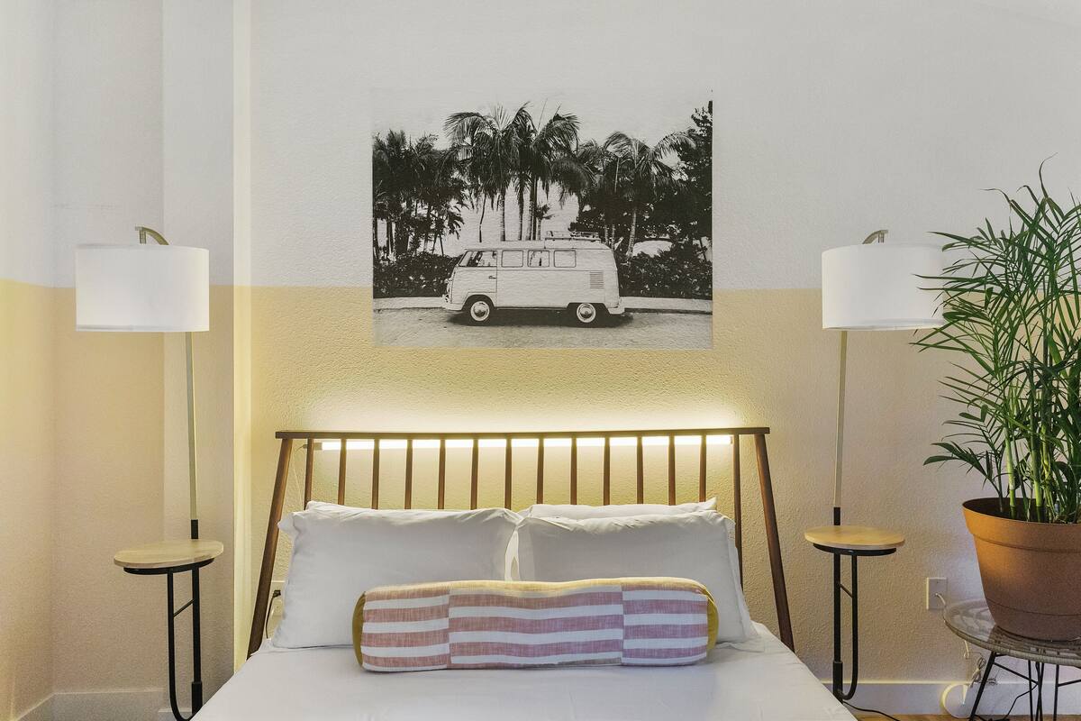 A neatly arranged hotel room with a bed, two bedside lamps, a wall-mounted picture, and a potted plant.