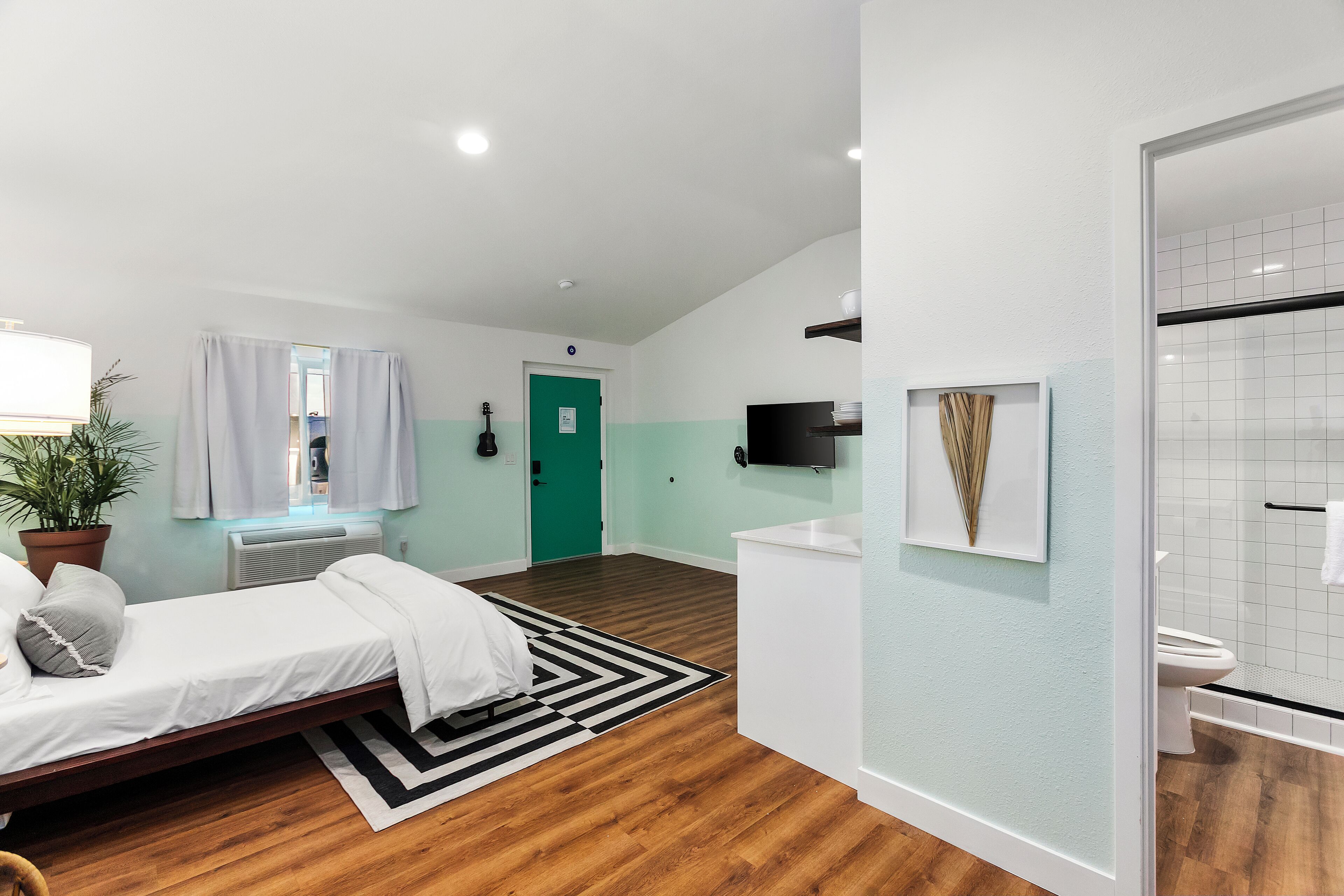 A modern room with a bed, a TV mounted on the wall, a green door, and a bathroom with a shower and toilet.