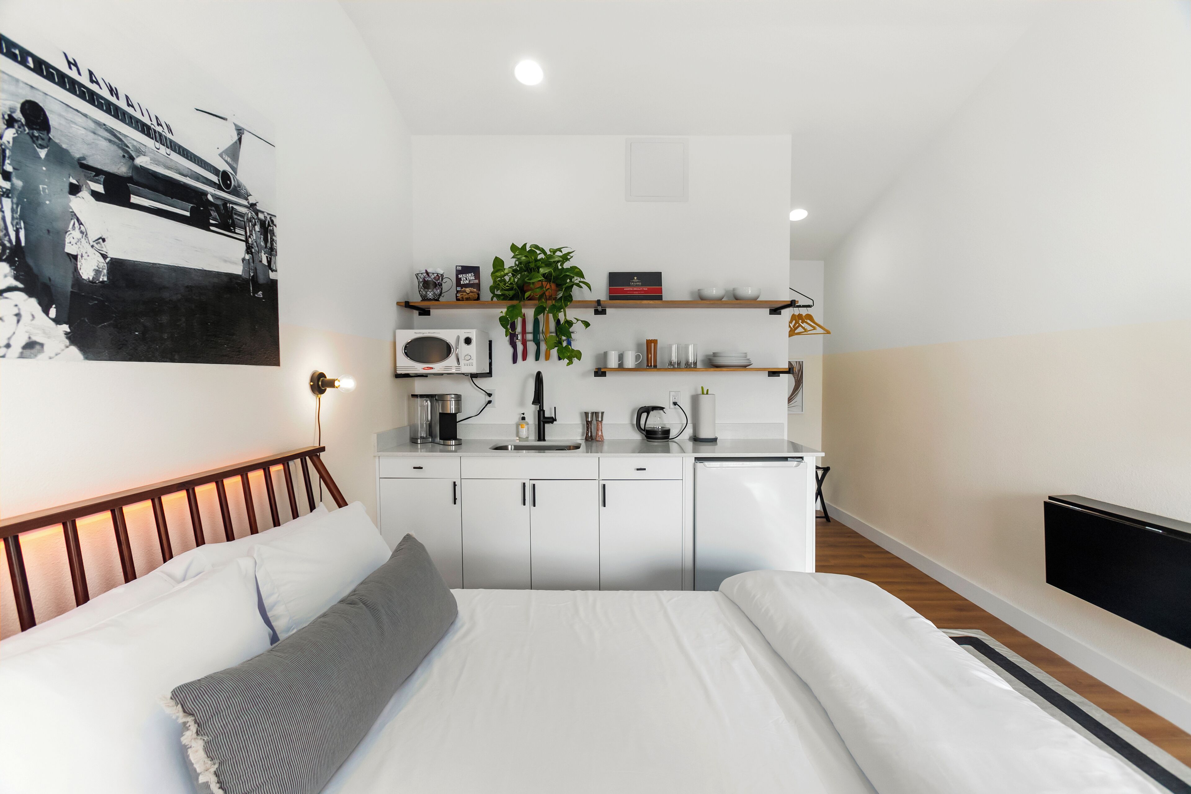 Una habitación de hotel compacta con una cama, kitchenette, televisión y un estante montado en la pared con varios objetos.