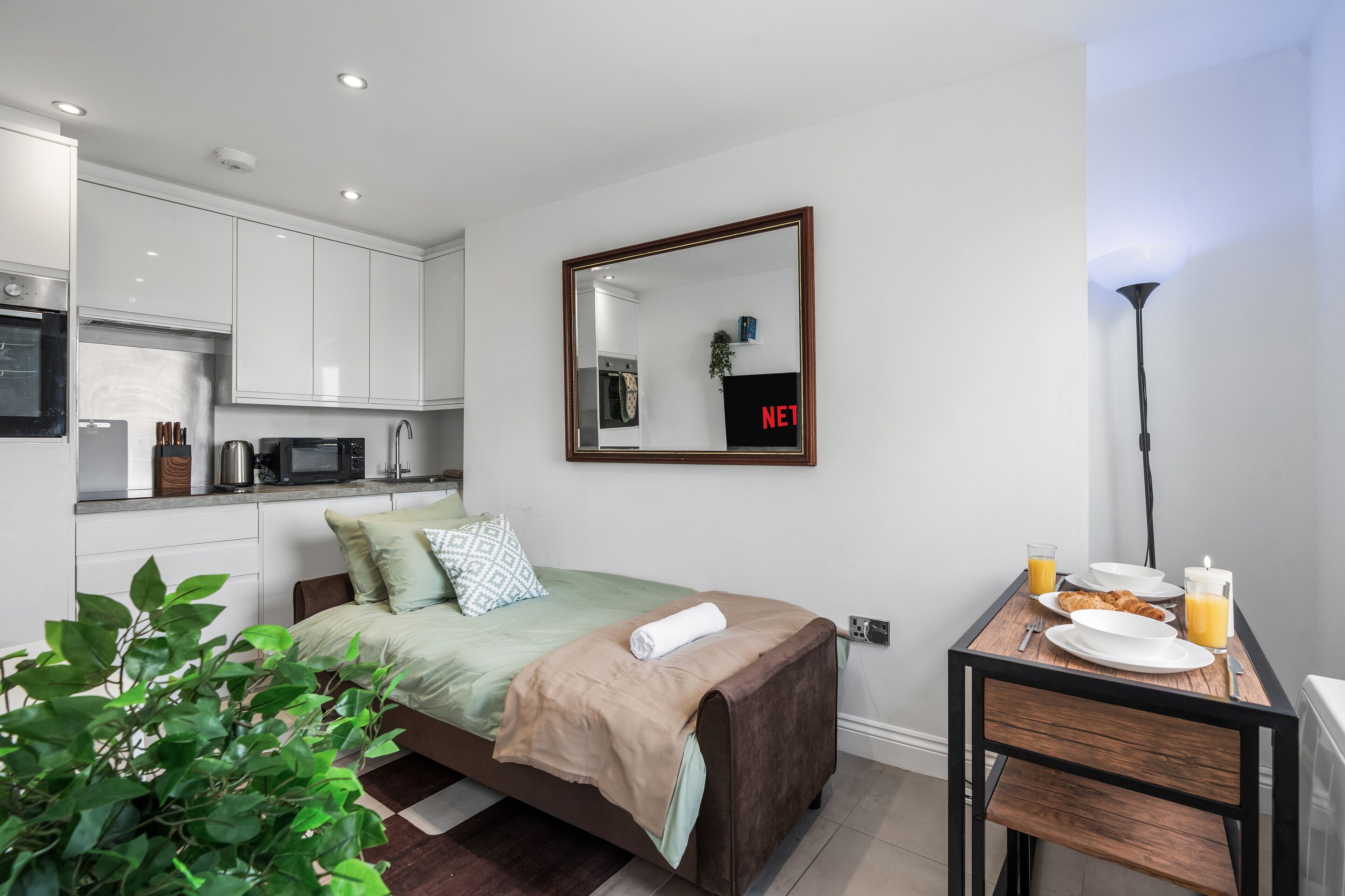 Une chambre d’hôtel compacte avec une kitchenette, un lit recouvert d’une courtepointe verte, une table de chevet avec des articles pour le petit-déjeuner et un miroir au mur.
