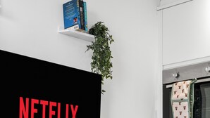 A modern living room with a large flat-screen TV displaying "NETFLIX" in red letters, a white table with a basket and remote, a kitchen area with an oven, and a small shelf with books and a plant.