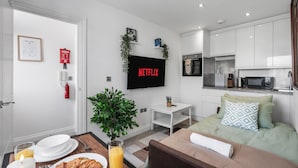 A modern hotel room with a kitchenette, a flat-screen TV, a dining area with breakfast items, and a bed with pillows.