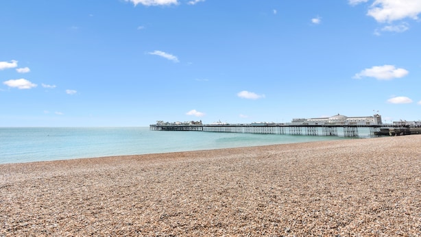 A pebbled beach with a pier extending into the sea and a building at the end of the pier.