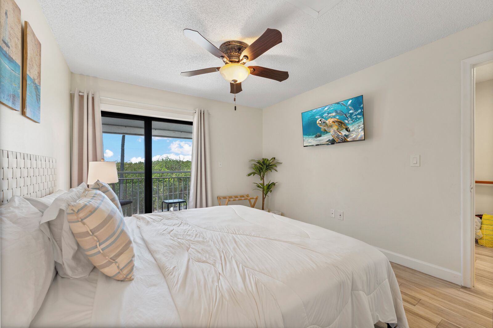 A bedroom with a bed, a ceiling fan, a window with curtains, a small table, and a plant.