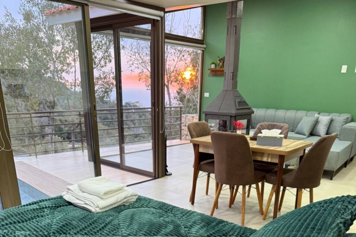 A bedroom with a large bed, a dining area with a table and chairs, a sofa, and a view of the outdoors through a sliding glass door.