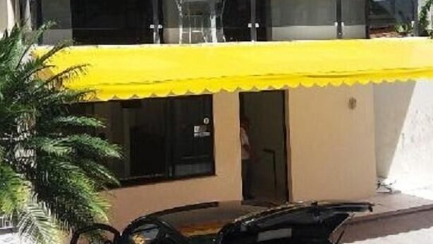 Un bâtiment à deux étages avec un auvent jaune, une voiture noire garée devant et une personne visible à travers la fenêtre.