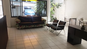 A room with a sofa, two chairs, a desk, and a window with a view of a parked car and greenery outside. - Pousada Vila Piatã (Salvador)