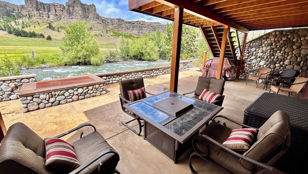 An outdoor patio with a stone wall, a glass-top table, and chairs with cushions. - Stillwater River Front Montana Home (Nye)