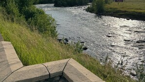 A river with a stone wall in the foreground and a house in the background. - Stillwater River Front Montana Home (Nye)