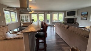 A modern kitchen with a central island, built-in cabinets, and a large window overlooking a green landscape. - Stillwater River Front Montana Home (Nye)