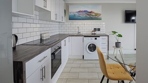 Une cuisine moderne avec des meubles blancs, un lave-linge encastré et un coin repas agrémenté d’une chaise jaune.